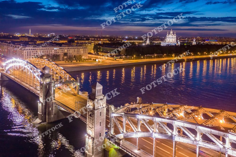 На переднем плане изображён Большеохтинский мост, переливающийся огнями ночного освещения. За мостом виднеется Смольный собор, добавляющий архитектурный акцент в ночной пейзаж Санкт-Петербурга. Отражение огней в воде Невы и спокойное ночное небо подчёркивают красоту и атмосферу города.